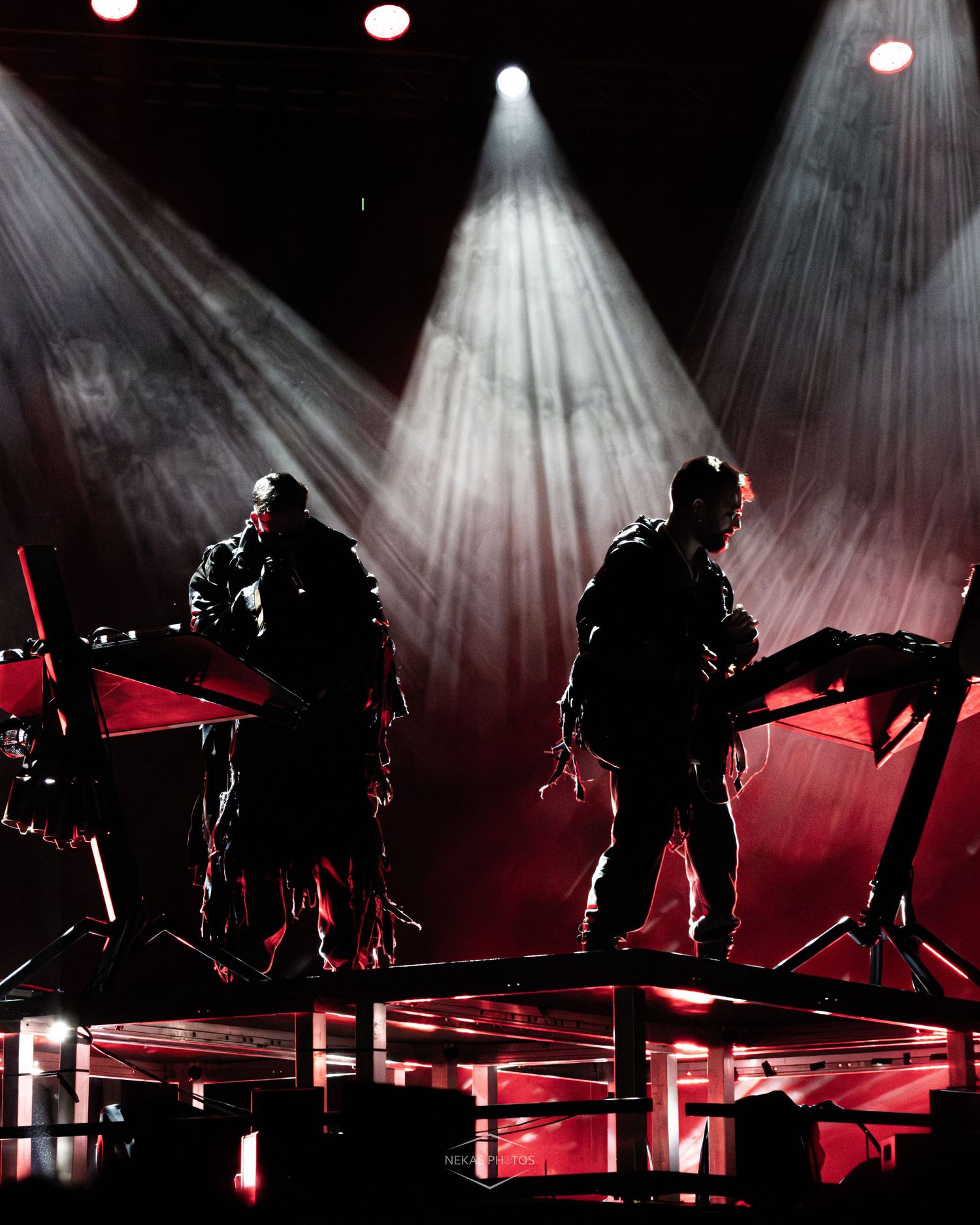 Two silhouetted musicians perform on a dramatic stage with red and white spotlights illuminating keyboards and equipment.