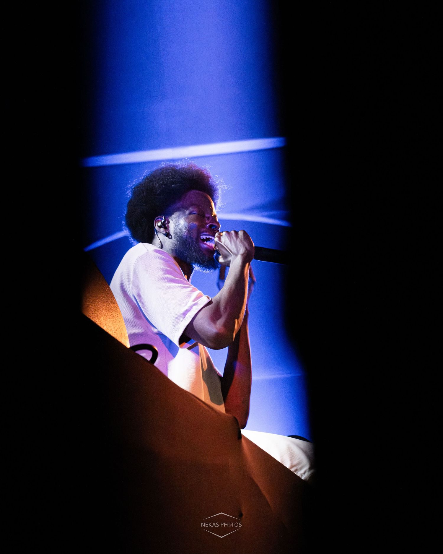 A young Black male performer with an afro sings into a microphone on a dramatic blue-lit concert stage.