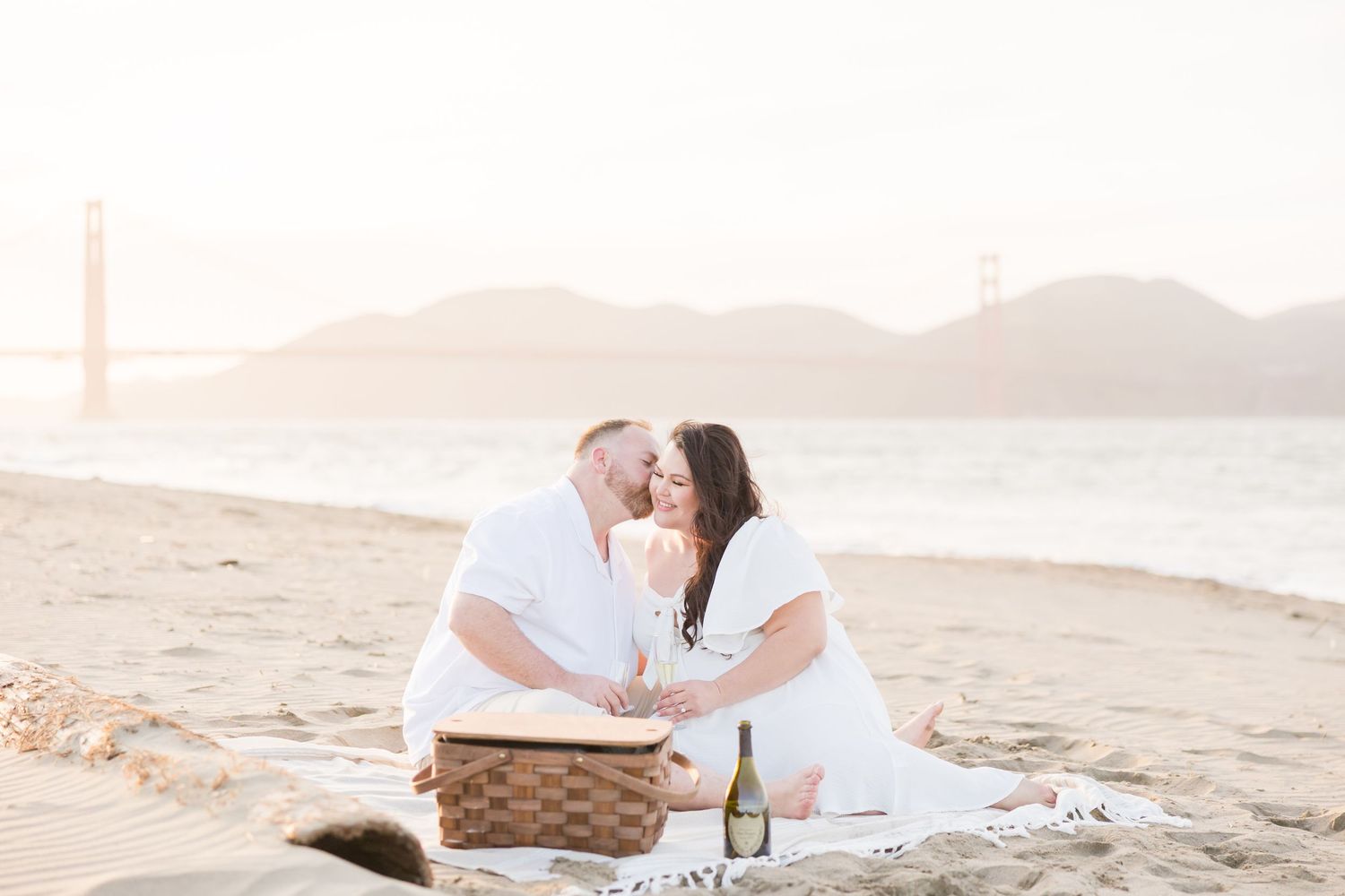 Couple embracing barefoot on a beach with the Golden Gate Bridge in the misty background.