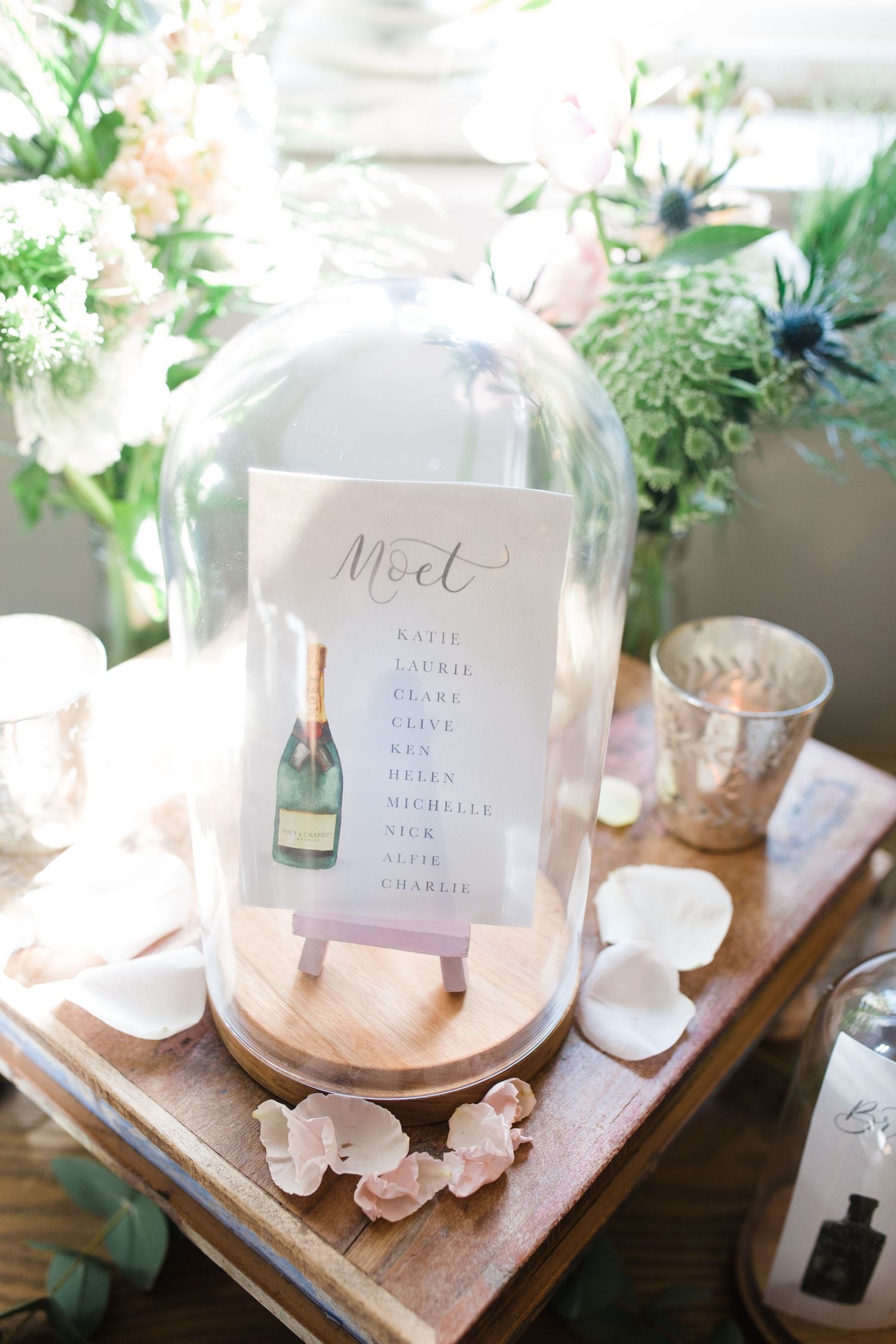 Elegant wedding seating chart under glass cloche dome on wooden base, surrounded by floral arrangements and rose petals.