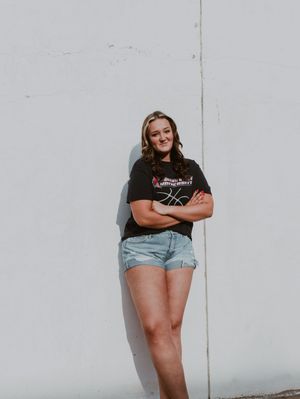 Young woman in black graphic tee and denim shorts poses confidently against white wall with arms crossed.