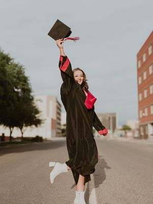 Excited graduate in black cap and gown jumping and raising her cap in the air on a city street.