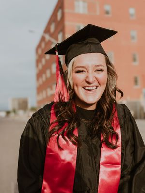 Happy female graduate smiling broadly in black cap and gown with red sash on a cloudy day outdoors.