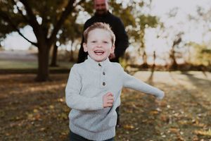 Happy red-haired toddler boy in gray sweater laughs joyfully outdoors in autumn park setting.