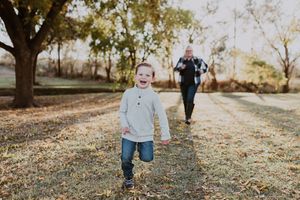 Laughing toddler runs toward camera on tree-lined path with woman following behind in autumn park.