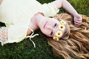 Young girl with long blonde hair lying on grass wearing yellow dress and daisy flower crown.