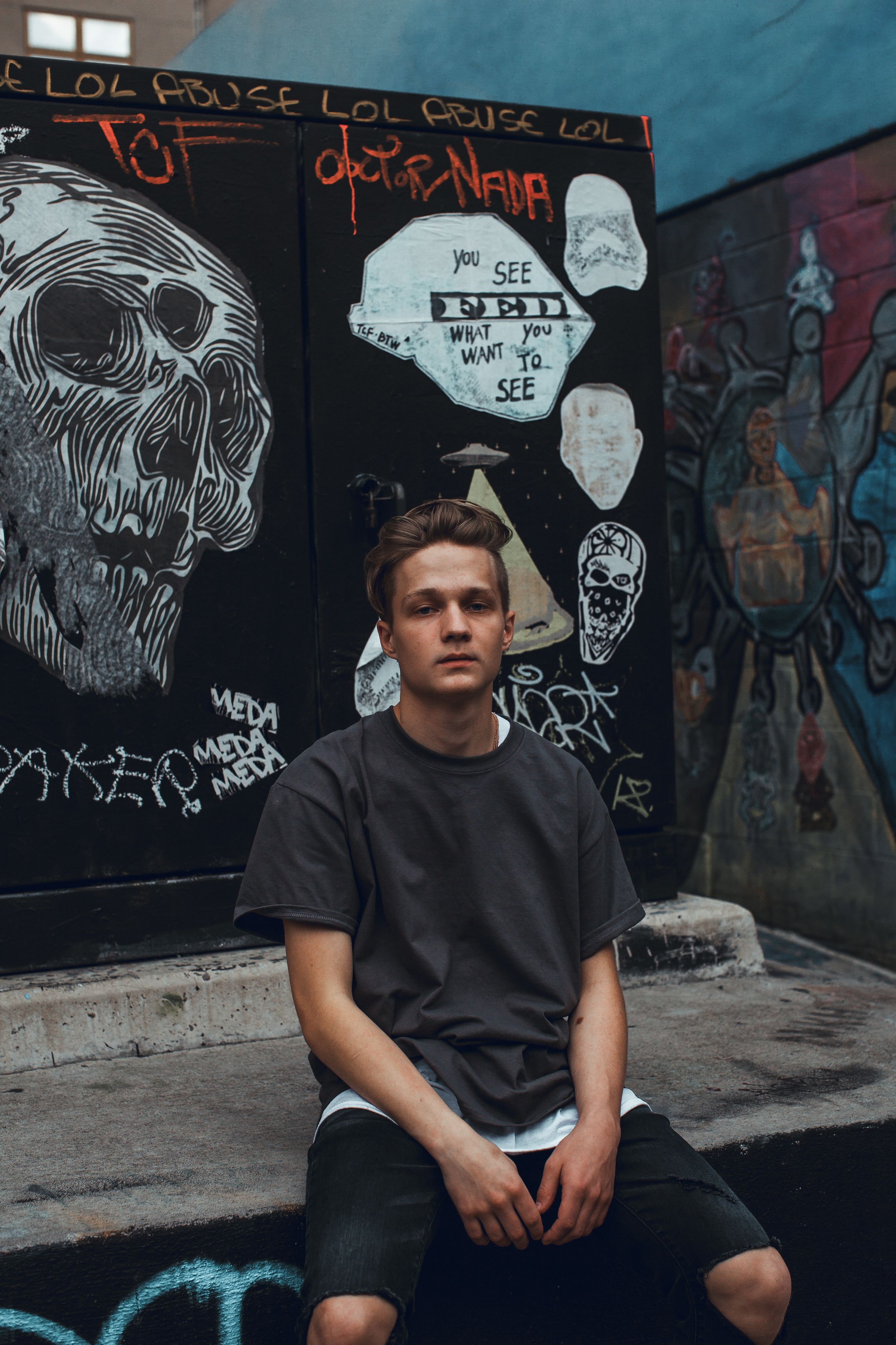 Young man in a dark t-shirt sitting in front of a graffiti-covered wall in an urban alleyway.