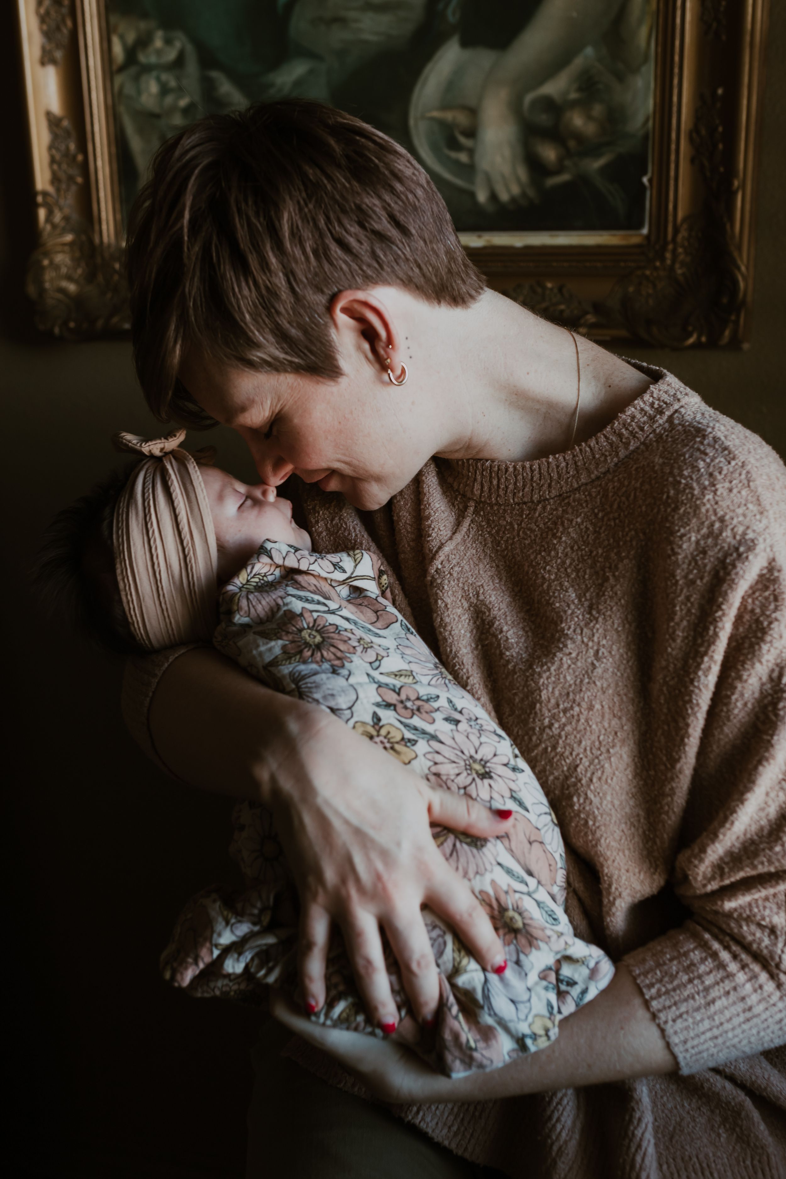 Mother with short hair lovingly nuzzling her swaddled newborn baby in dim lighting.