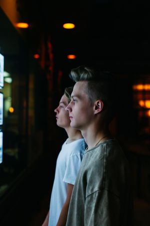 Two young men standing side by side in profile view in a dimly lit room with warm bokeh lights.