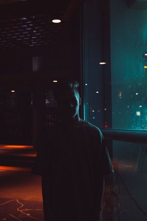 Silhouette of a person standing alone looking out a rain-covered window at night with colorful lights.