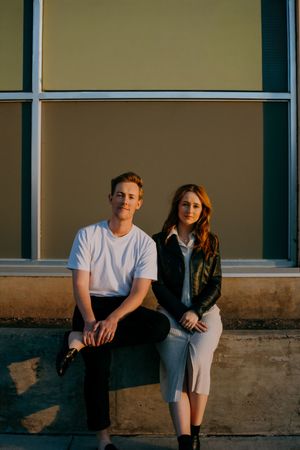 Young man and woman sitting together on a ledge outside a building in warm golden hour light.