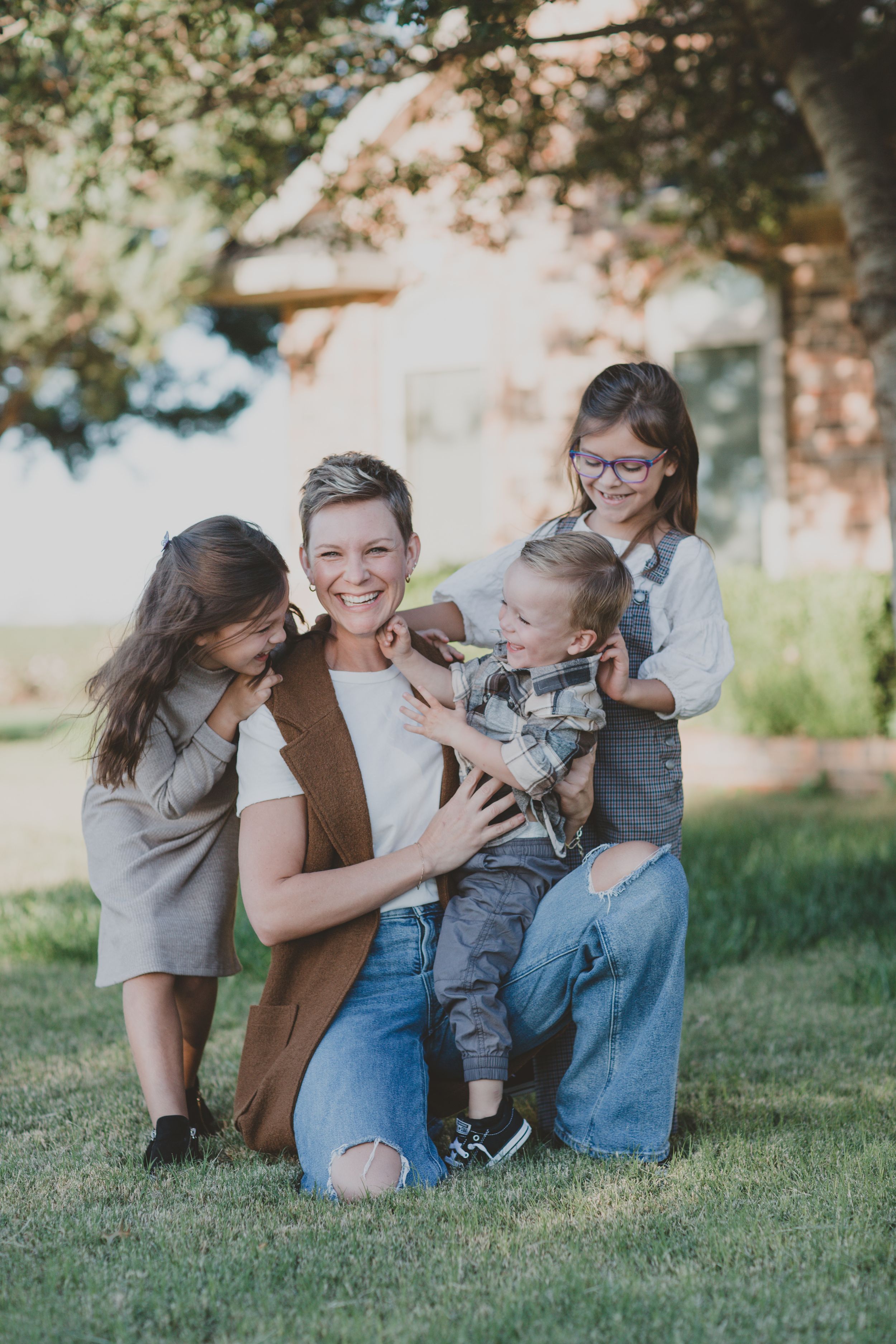 Happy mother kneeling on grass laughing with three young children in outdoor setting.