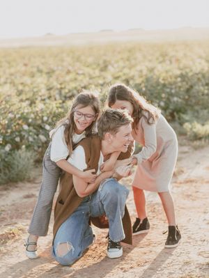 Parent crouching in cotton field being hugged by two laughing children at golden hour.