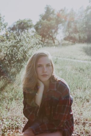 Young blonde woman sitting in a sunlit green field wearing a red plaid shirt with soft natural light.