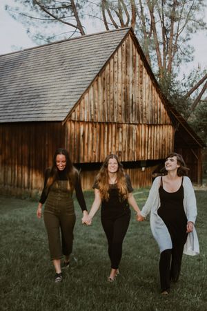Three young women holding hands and walking together in front of a rustic wooden barn outdoors.