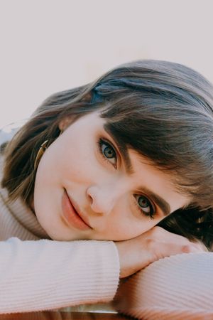 Close-up portrait of a young woman with blue eyes resting her chin on her hands with a soft smile.