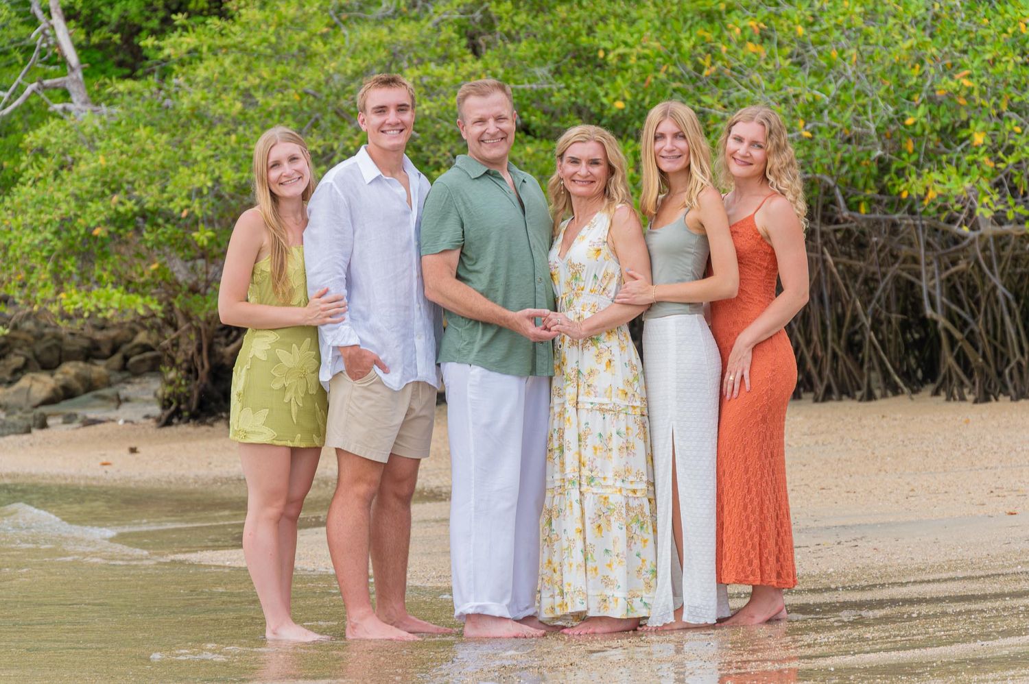 Family of five smiling together on a tropical beach with lush green trees in the background.