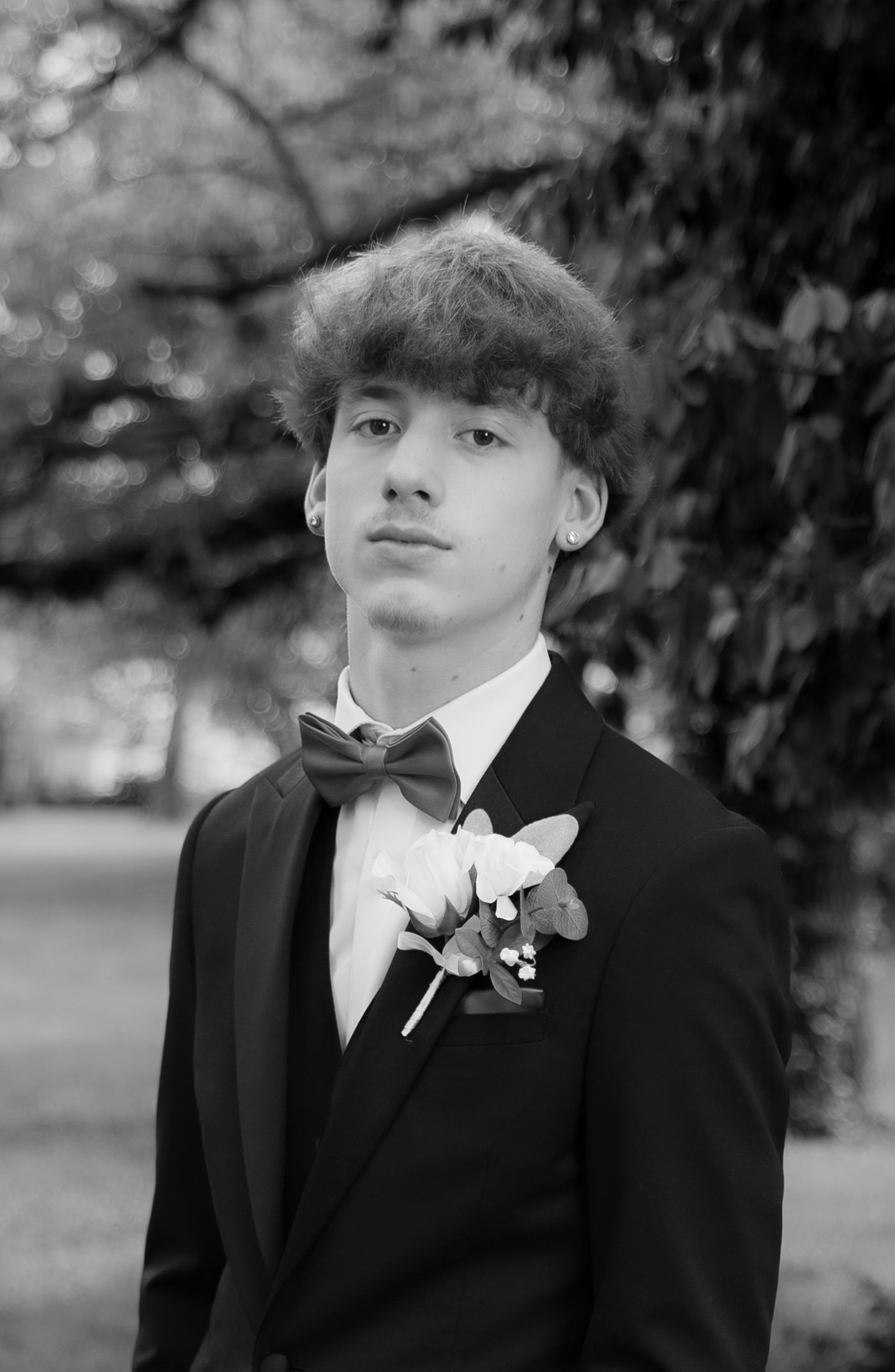 Young man in black tuxedo with bow tie and boutonniere poses outdoors in black and white formal photo.