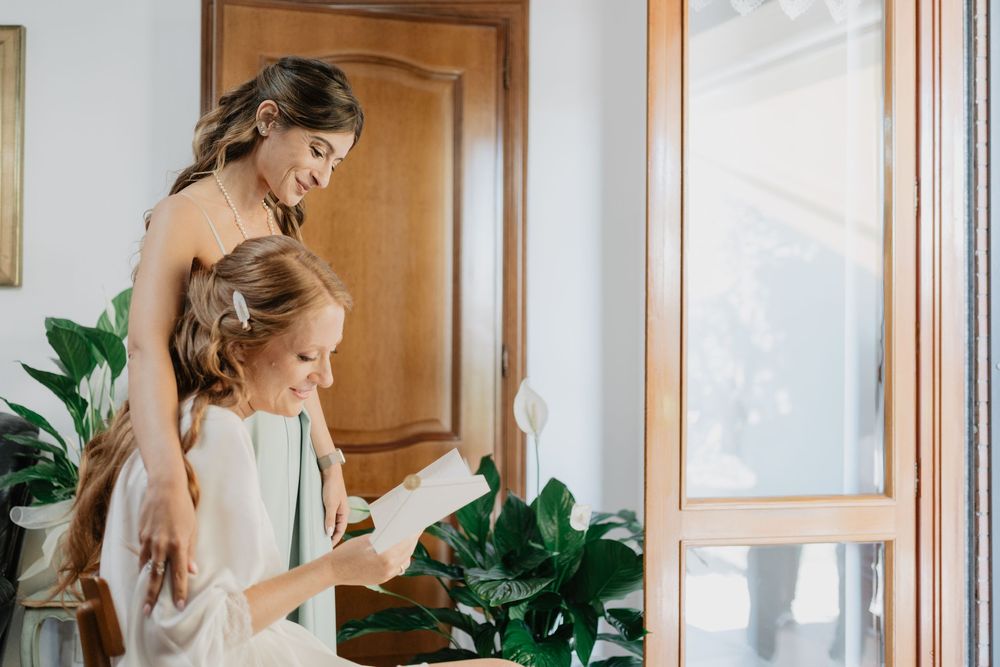 Two women share a joyful moment reading together near a sunlit window with lush green plants.