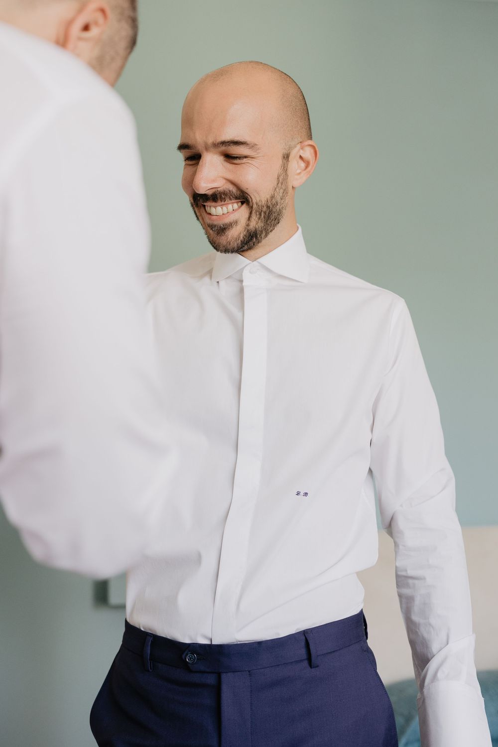 Smiling bald groom in white dress shirt and navy trousers getting ready with groomsman before wedding.