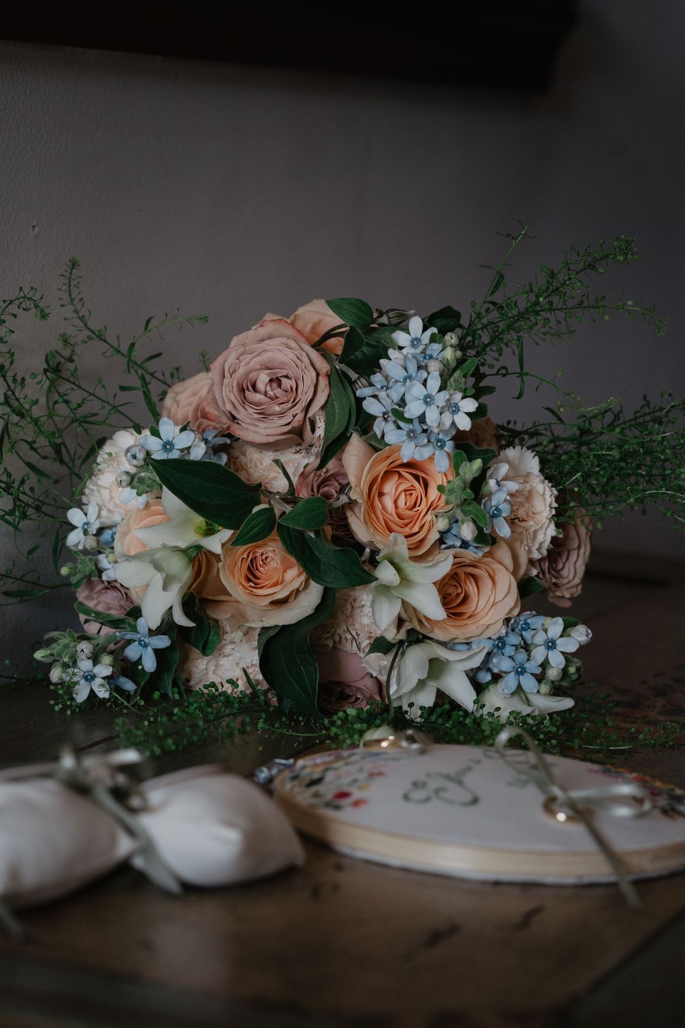 Elegant wedding bouquet with peach roses, blue hydrangeas, and greenery on a table with ring pillow.