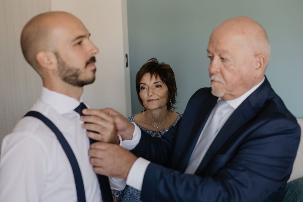 Elderly man helps groom adjust his tie while woman watches during wedding day preparations.