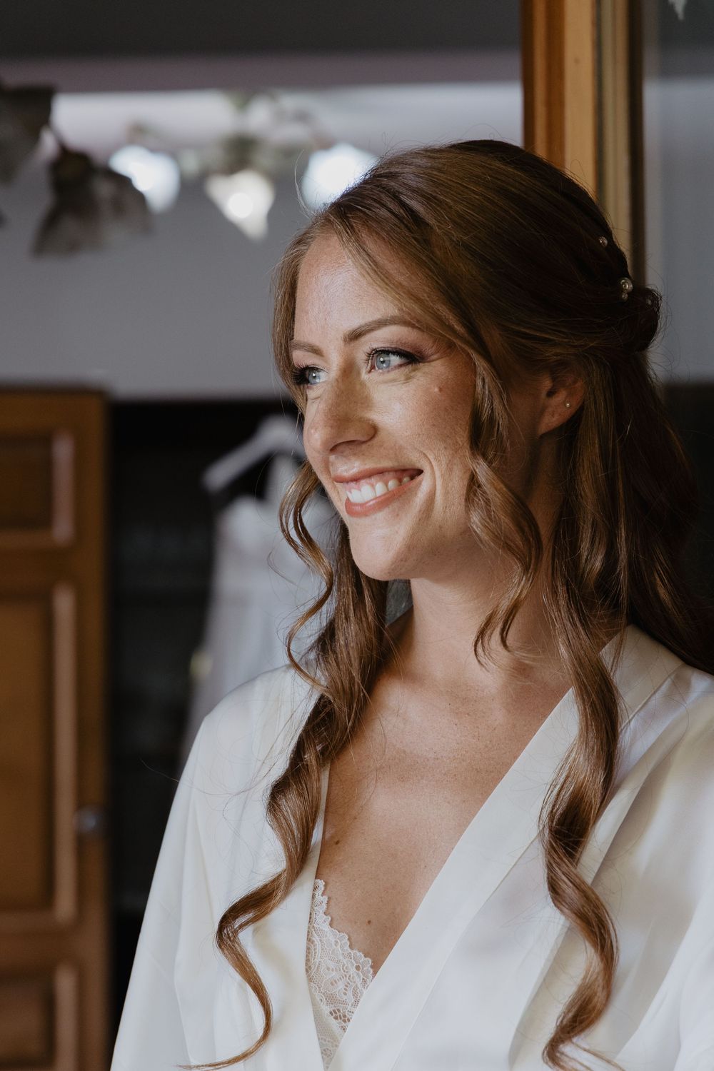 Smiling bride with wavy auburn hair in a white robe during wedding morning preparations.