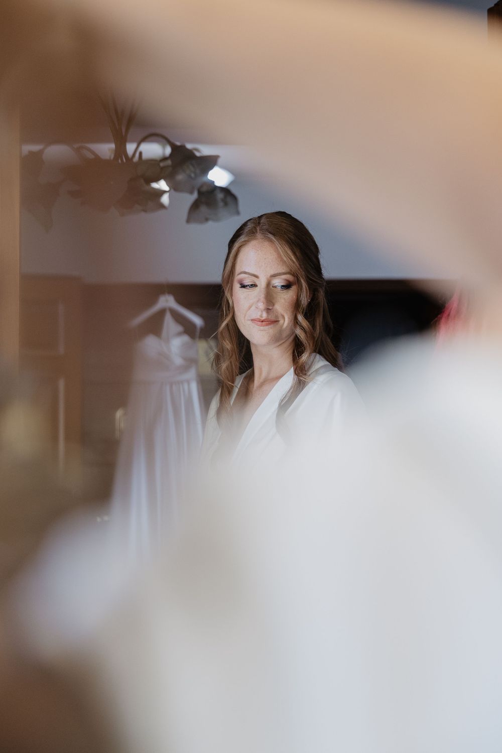 Beautiful bride in white robe seen through a mirror reflection during wedding day preparations.