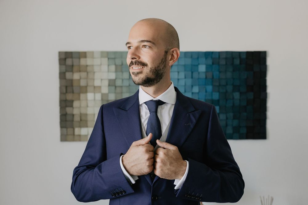 Elegant bald groom in navy blue suit adjusting jacket before wedding ceremony indoors.