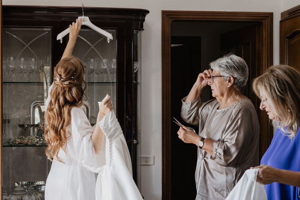 Bride in white robe holding wedding dress on hanger while older women assist during preparations.