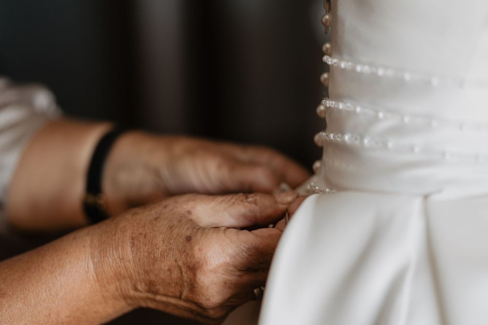 Hands fastening pearl buttons on the back of a white wedding gown.