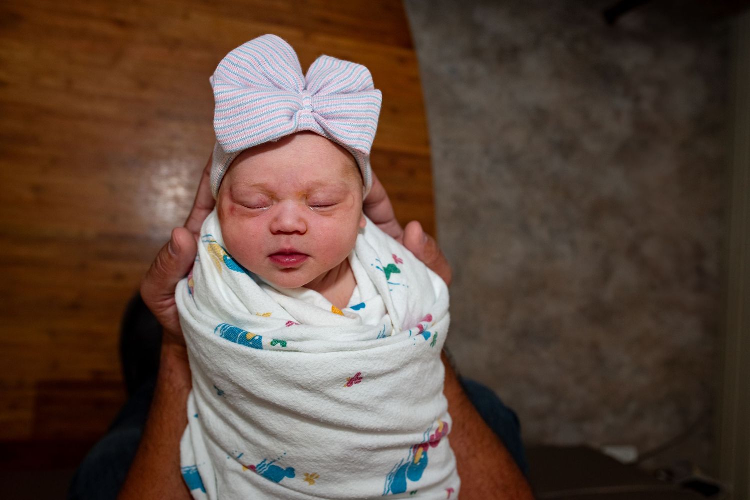 A sleeping newborn baby swaddled in a white patterned blanket and wearing a white bow headband.