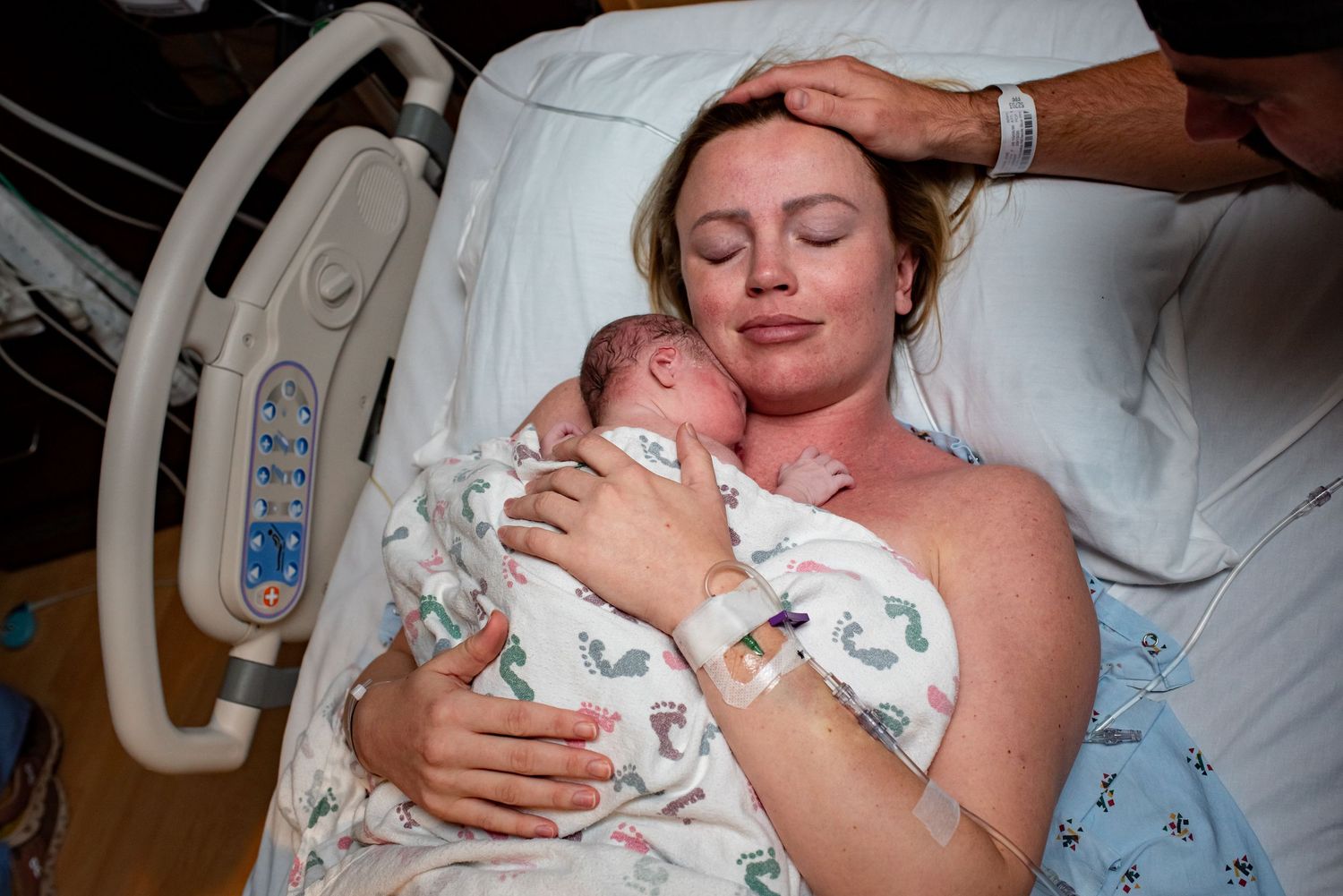 A tender moment in a hospital room as a parent cradles their newborn baby wrapped in a floral print blanket.