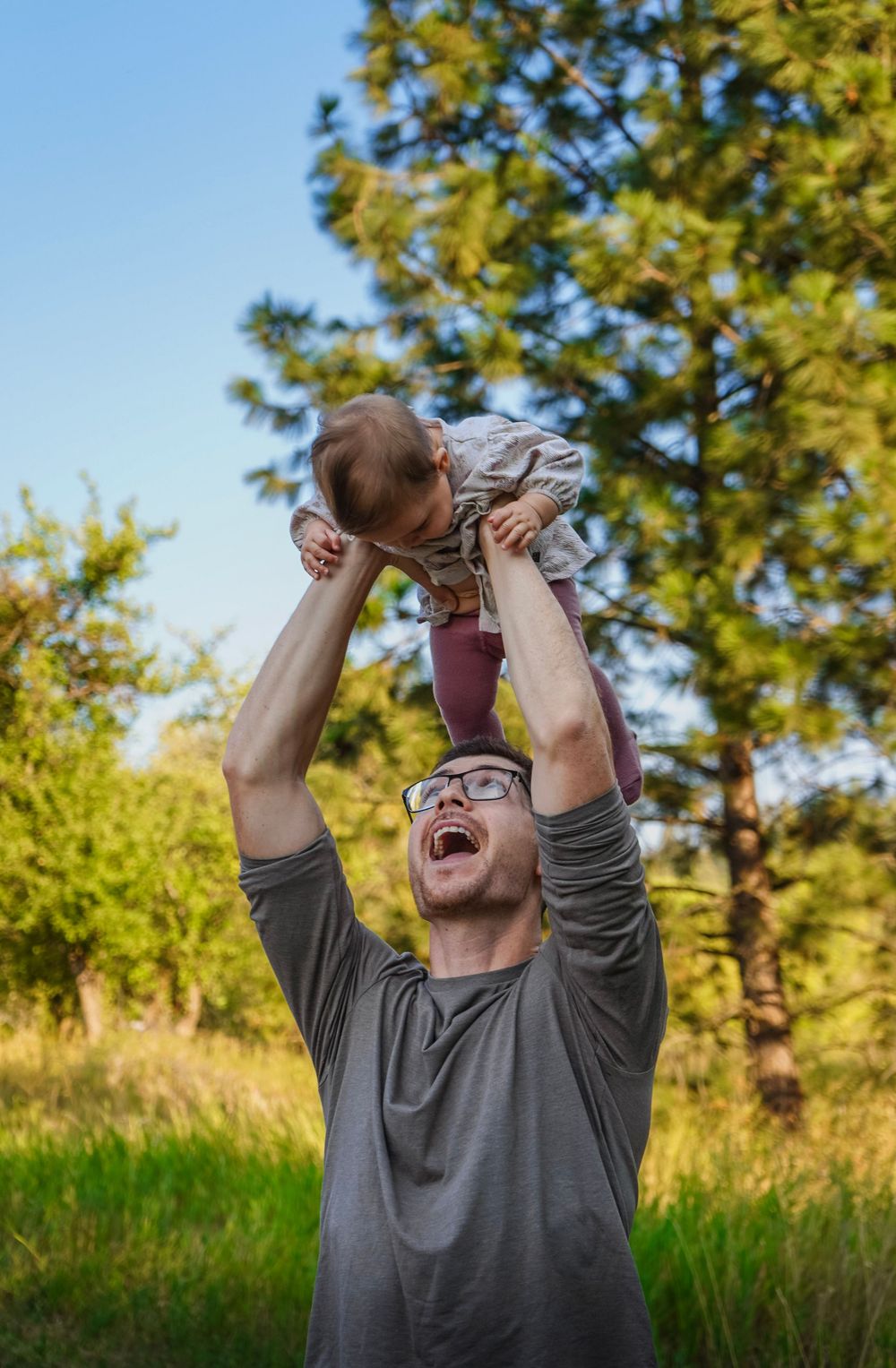 A joyful father lifts his baby overhead outdoors, both smiling, surrounded by lush green trees on a sunny day.