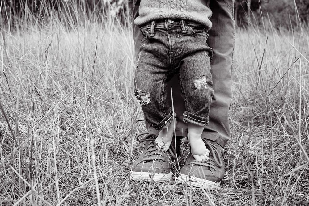 Black and white photo of a child standing between an adult's legs in a grassy field, both wearing jeans and sneakers.