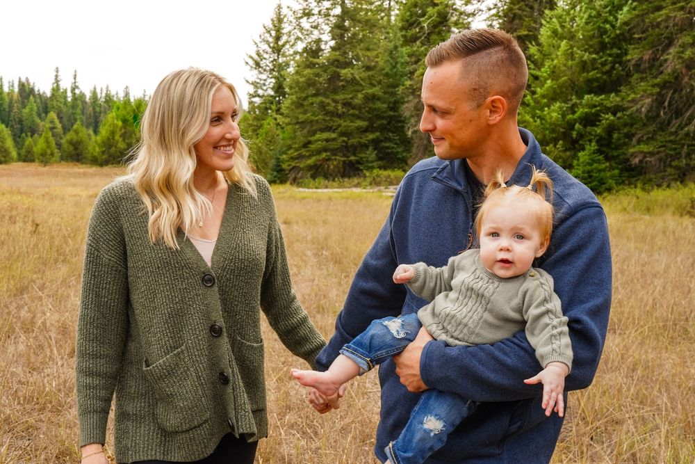 Young family with blonde mother, father, and baby in arms smiling in golden meadow with pine trees.
