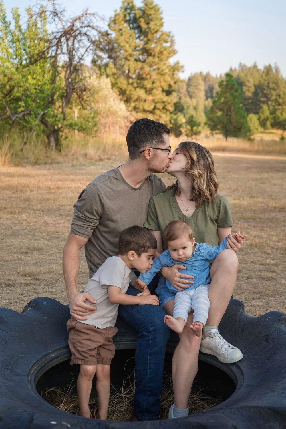 A family of four poses outdoors on a large tire, parents kissing while holding their two young sons in a sunny field.