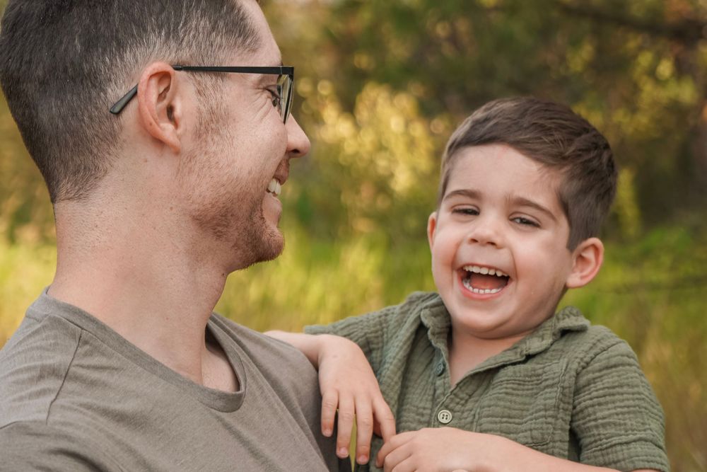 A father and young son share a joyful laugh outdoors, with the boy smiling broadly in a green shirt.