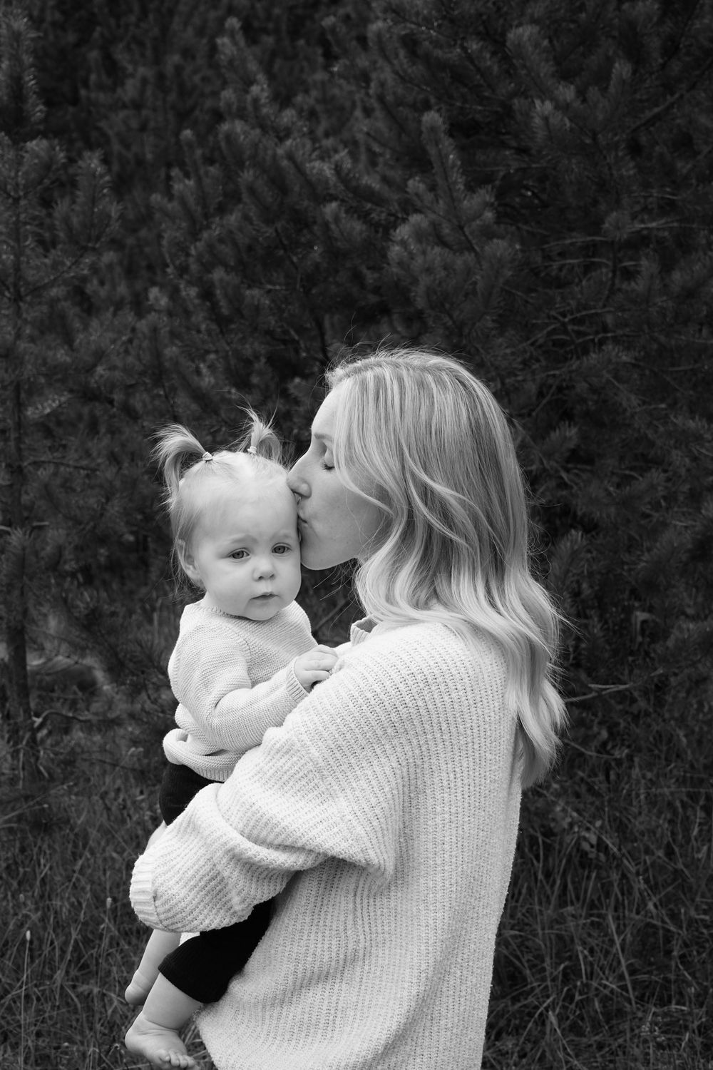 Black and white photo of a blonde woman kissing a toddler girl she holds in her arms outdoors.