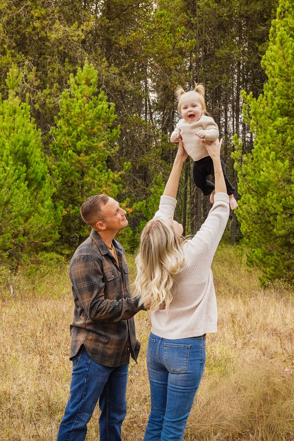 A couple plays with their laughing baby outdoors, lifting the child high in a forest meadow during a family photo session.