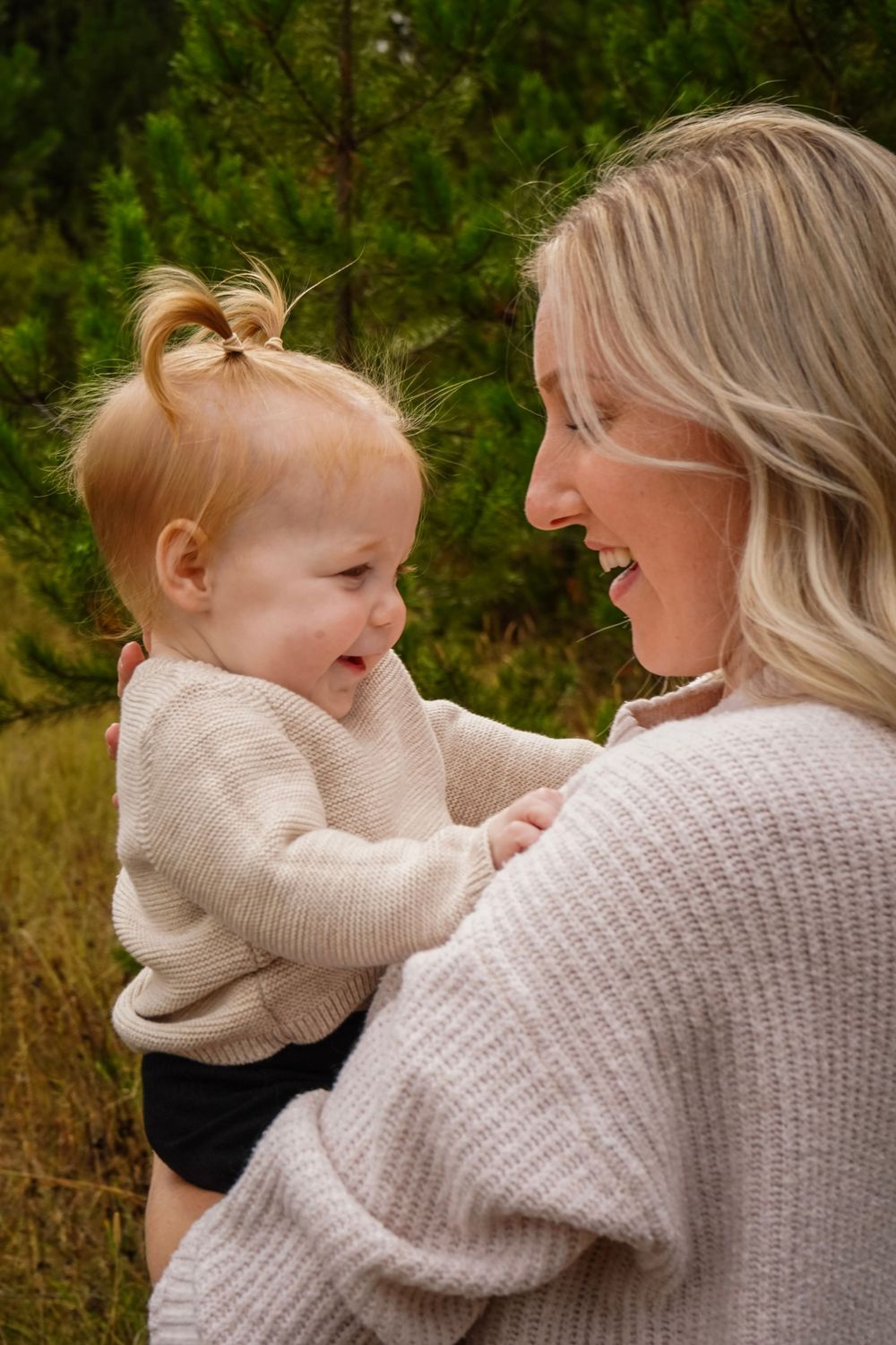 Blonde mother smiling and holding her baby girl outdoors near evergreen trees in matching cream sweaters.