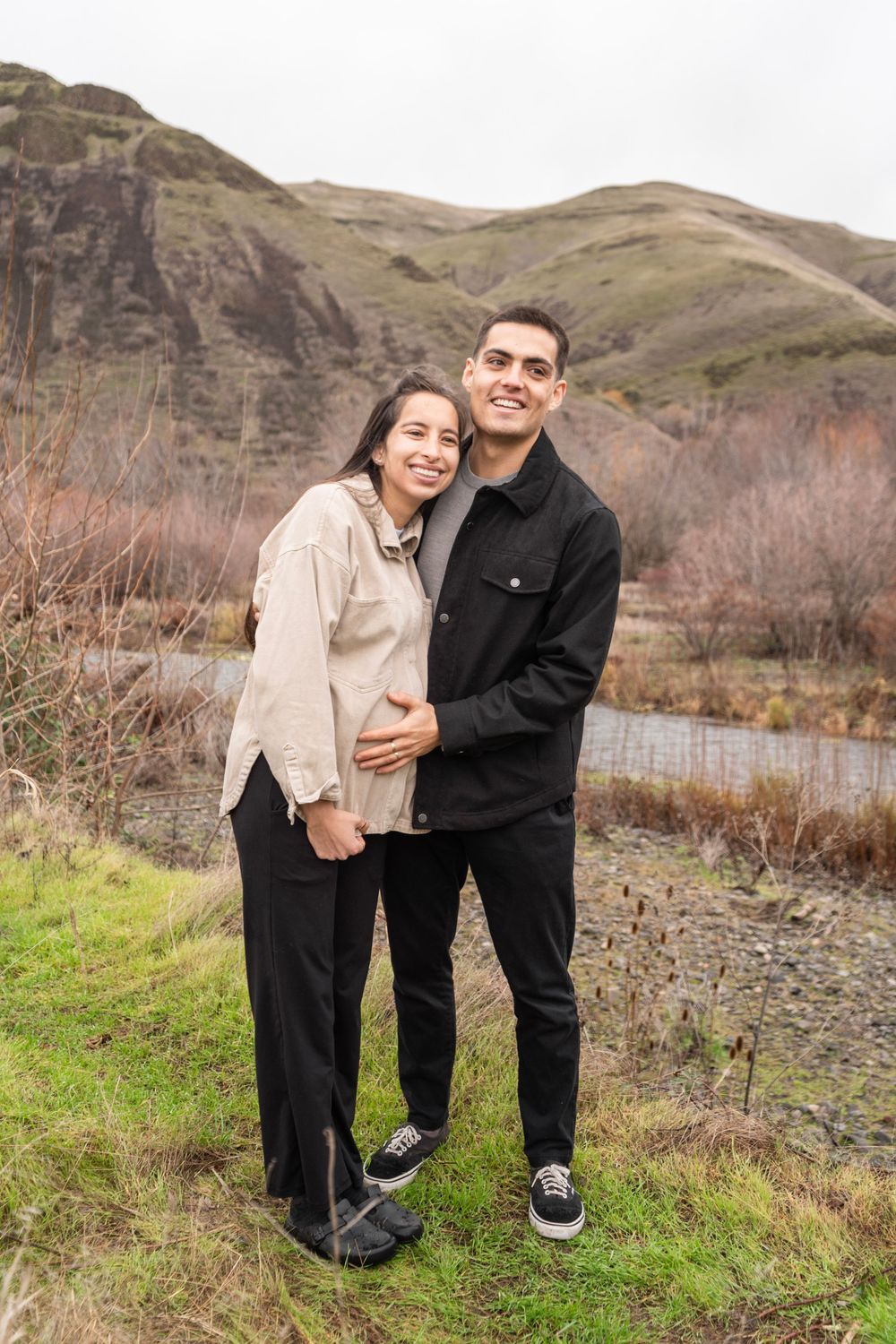Smiling couple embracing outdoors with scenic hills and river in the background.