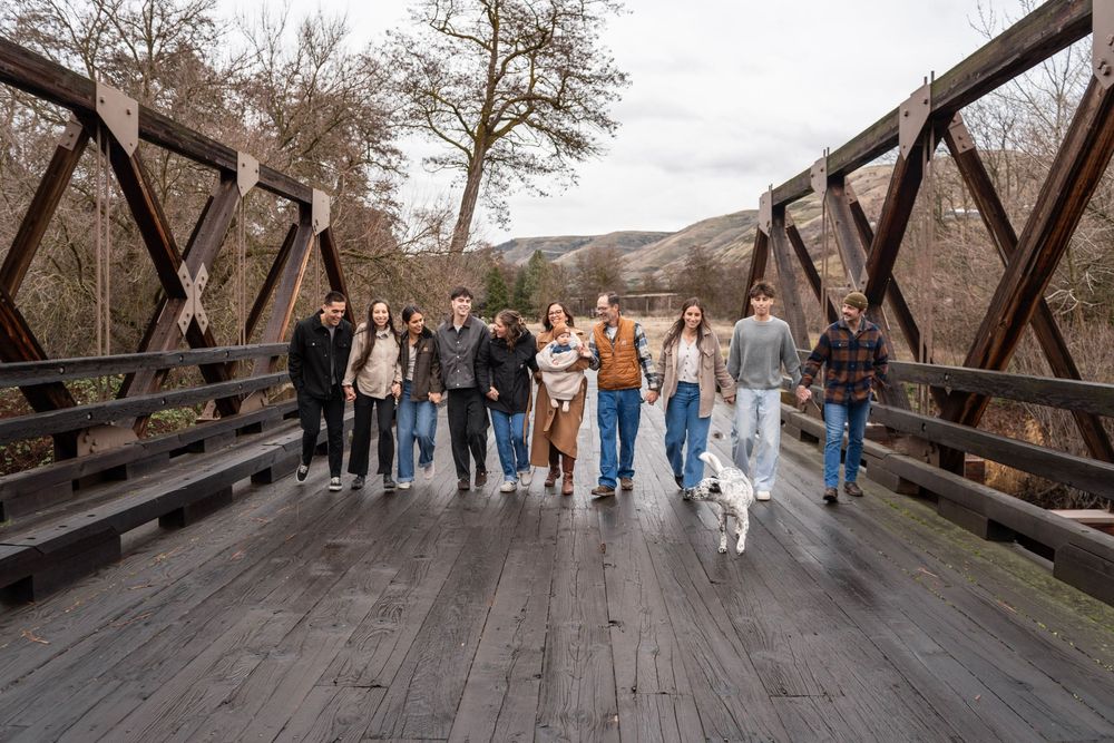 Group of friends walking a white dog across a wooden bridge surrounded by trees in a scenic outdoor setting.