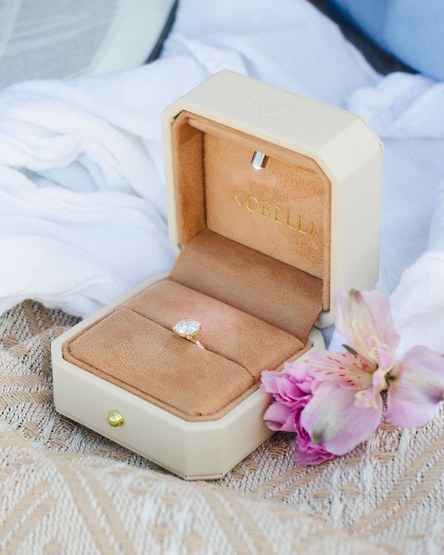 Diamond engagement ring in an open cream velvet Norella jewelry box with pink flowers on a lace surface.
