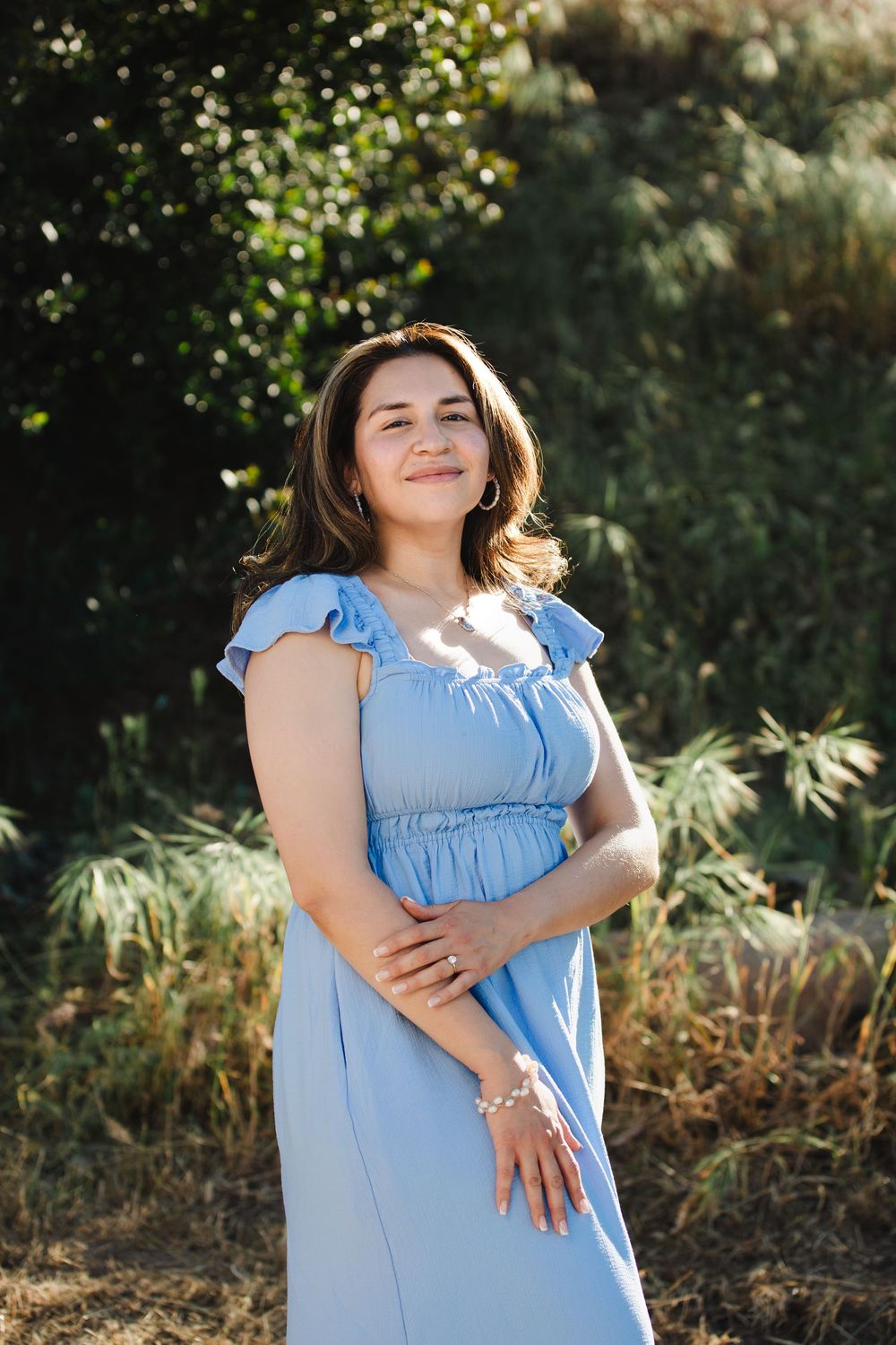 Woman in light blue ruffle dress smiling outdoors in golden sunlight with green foliage background.