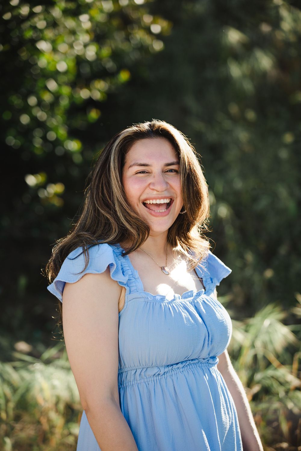 Laughing young woman with brown hair wearing a light blue ruffle dress outdoors in a sunlit garden.