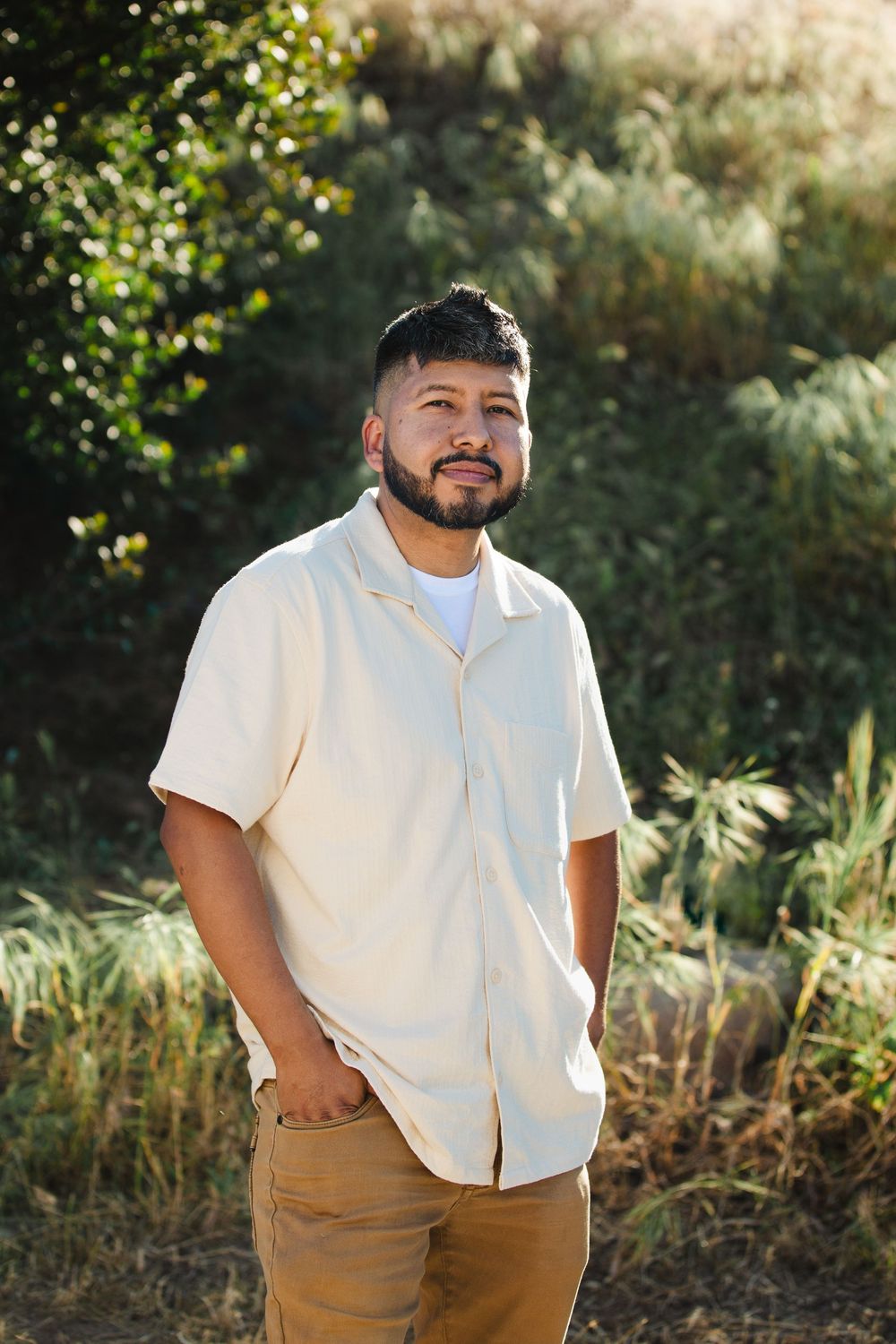 Man in beige shirt standing outdoors with lush green foliage in the background on a sunny day.