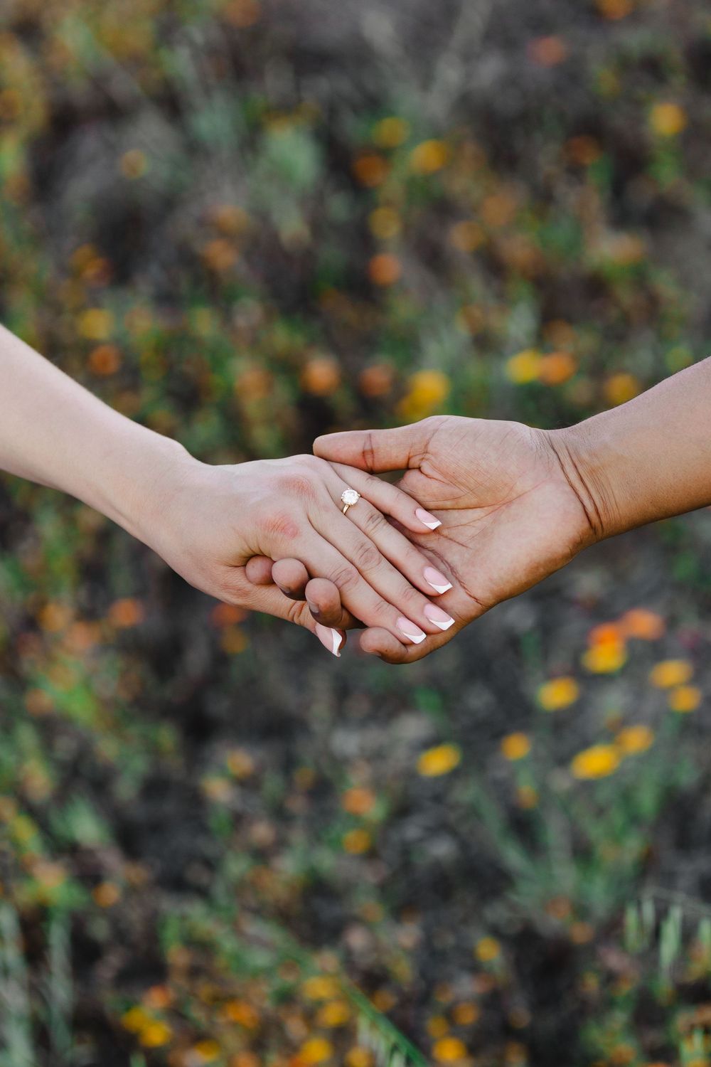 Two people holding hands outdoors in a flower field, one wearing a gold engagement ring with white manicured nails.