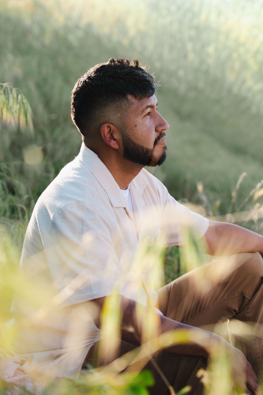 Bearded Hispanic man in white shirt sitting thoughtfully in lush green grass field in natural sunlight.
