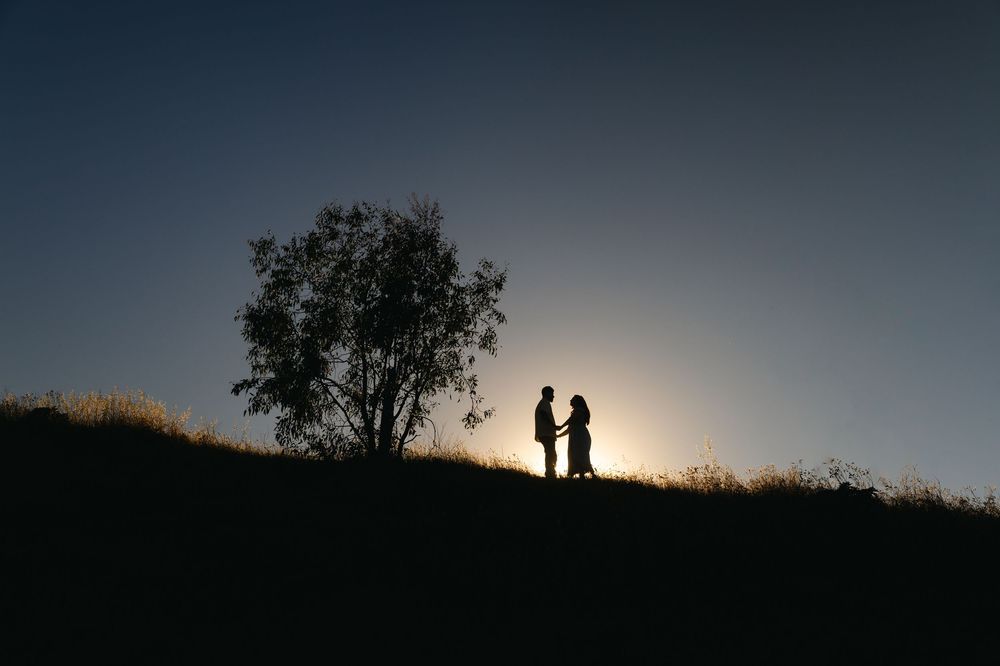 Romantic couple silhouette standing near a tree on a hill at sunset with glowing sky backdrop.